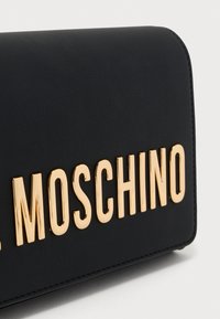 Bolso de mano de cuero negro con acabado texturizado, con letras doradas que dicen "MOSCHINO" de manera prominente en el frente. Diseño limpio y rectangular.