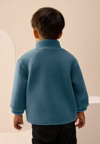 Kék, bolyhos példányú fleece dzseki magas gallérral, laza szabással és ejtett vállakkal, hátulról nézve.