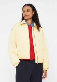 Dzeltena pūkaina jaka ar rāvējslēdzēju priekšā, ribotām aprocēm un logotipa detaļu. Apvienota ar sarkanu adītu džemperi un zilu krekla blūzi.