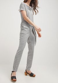 Jumpsuit a motivo bianco e nero con scollo a barca e vita legata. Abbinato a sandali con rialzo neri, con le dita dei piedi scoperte e tacchi in legno.