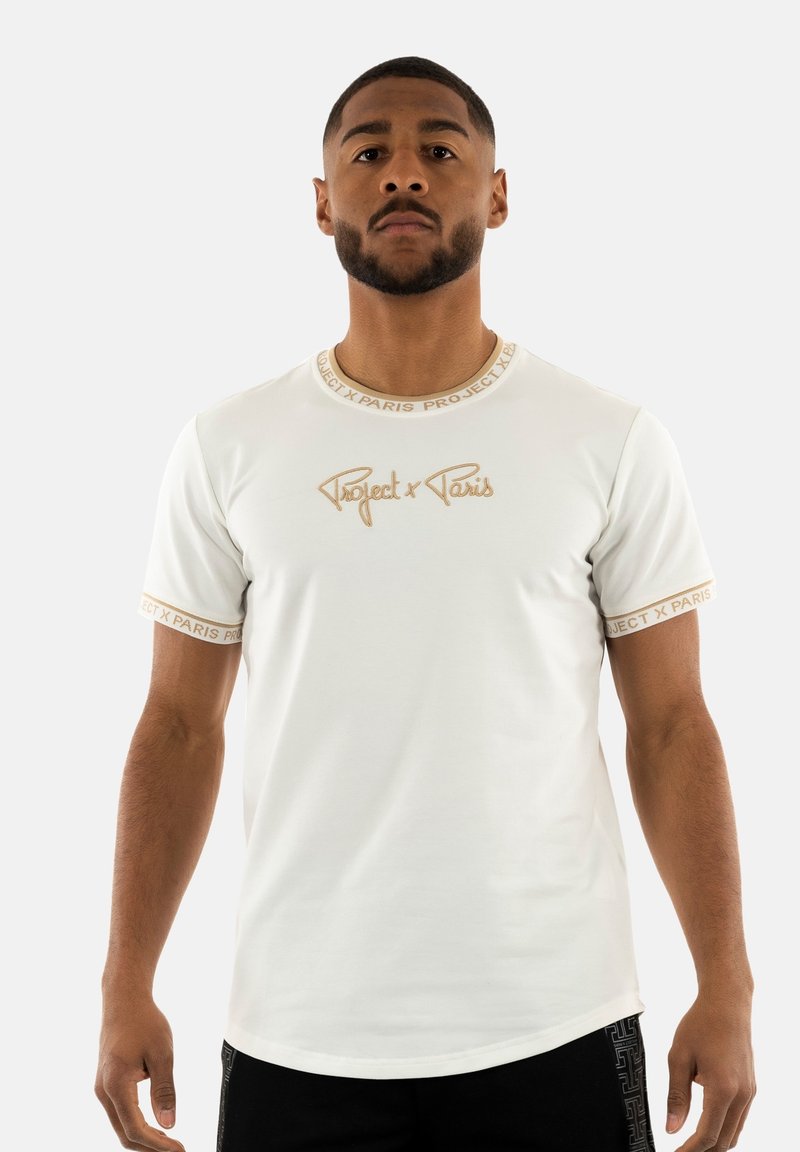 Camiseta blanca de manga corta con cuello redondo, logo bordado "Project x Paris" en dorado y ribete dorado contrastante en las mangas.