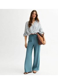 Randig skjorta med knutar i midjan; lösa, turkosa byxor med bred benvidd; brun handväska med mjuk textur; sandaler med tunna remmar.