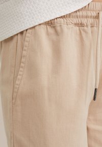 Beiga joggers med dragsko i midjan, sidofickor och en mjuk bomullsblandningstextur. Accenter inkluderar en silverfärgad dragkedjedetalj.