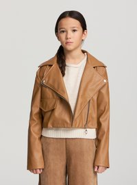 Giacca corta marrone in similpelle con colletto a revers, chiusura con cerniera e dettagli con tasche laterali, indossata sopra un maglione beige e pantaloni marroni.