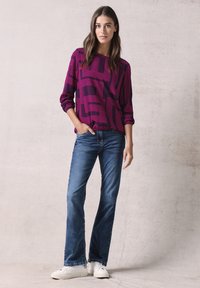 Camisetta viola a maniche lunghe con motivo geometrico rosa, abbinata a jeans a zampa d'elefante blu chiaro e sneaker bianche, su uno sfondo neutro.