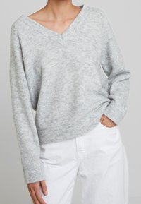 Hellgrauer, strukturierter Pullover mit V-Ausschnitt und gerippten Bündchen. Trägt man über weißen Hosen, die eine lockere Passform und weichen Stoff zeigen.