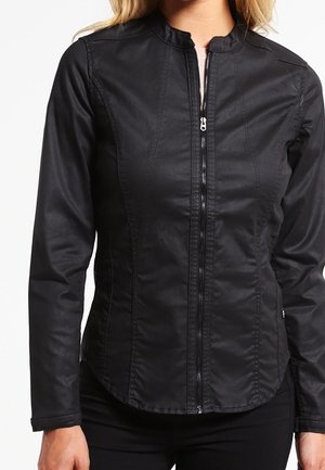 Femme portant une veste noire ajustée avec fermeture éclair frontale, col haut et manches longues, vue des épaules jusqu'aux hanches.