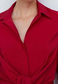 Chemise rouge avec col pointu, fermeture à boutons et détail noué à la taille. Tissu lisse de couleur unie avec coupe ajustée.