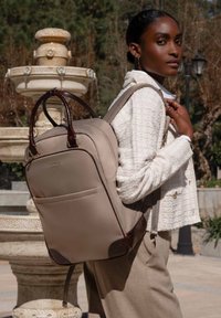 Sac à dos beige avec des accents en cuir marron, deux poignées supérieures, une poche avant et des sangles rembourrées ; texture lisse et design structuré.