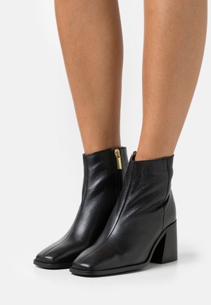 Bottines en cuir noir avec bout carré, talon bloc haut et détail de fermeture éclair dorée. Texture lisse avec un design minimaliste.