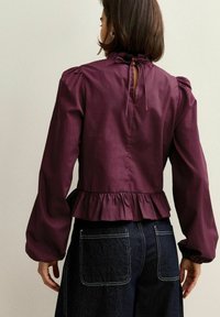 Burgundy blus med lång ärm, puffiga axlar, volangkant och nyckelhål bak. Kombinerad med mörk denimbyxor med kontrasterande sömmar.