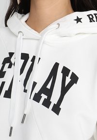 Bílá mikina s kapucí, na které je velkými černými písmeny nápis "REPLAY", s šňůrkami a hvězdicovým vzorem na podšívce kapuce.