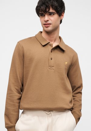 Bruine polo met lange mouwen en kraag, voorzien van twee knopen en een klein geel logo. Het materiaal lijkt textuur te hebben en is gecombineerd met beige broek.