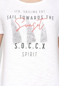 Camiseta de algodón blanca con un estampado gráfico que incluye el texto "Navega hacia el atardecer" y formas de veleros grises. Texto en negro y rojo.