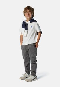 Weißes Polohemd mit blauem Logo, graue Cargo-Hosen und helle Sneakers. Dunkelblauer Pullover um den Hals gebunden. Junge steht lächelnd.