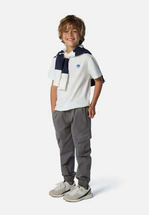 Weißes Polohemd mit blauem Logo, graue Cargo-Hosen und helle Sneakers. Dunkelblauer Pullover um den Hals gebunden. Junge steht lächelnd.