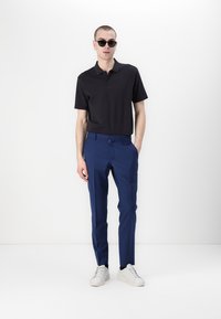 Schwarzes Polohemd, maßgeschneiderte blaue Hose, weiße Sneakers. Accessoires umfassen dunkle Sonnenbrillen und einen Metallohrring. Glatte Stofftextur.