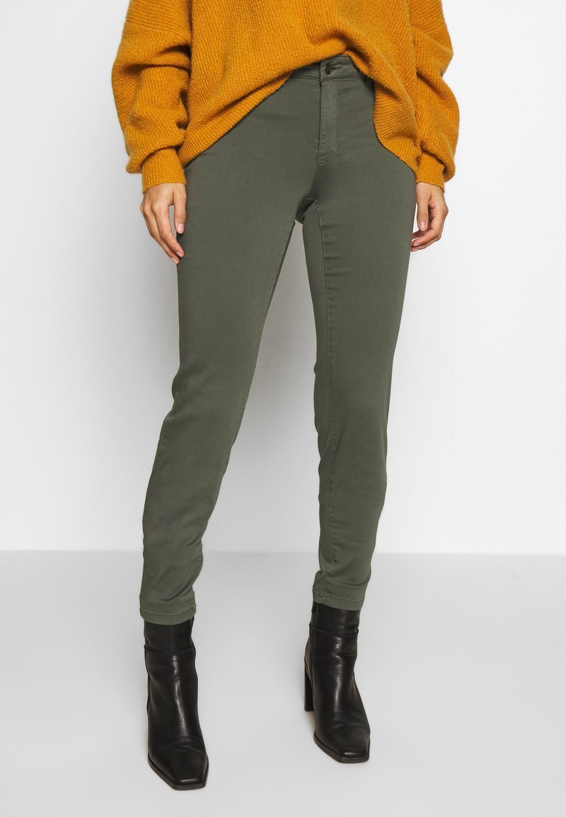 Personne portant un pantalon slim vert olive, un pull en tricot moutarde et des bottines à talons noires, debout devant un fond clair uni.
