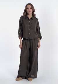 Completo in due pezzi in lino di colore marrone scuro; presenta una camicia corta con bottoni e tasca e pantaloni a gamba larga con vita elastica.
