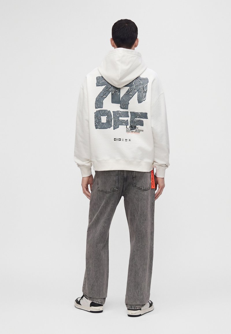 Homme portant un sweat à capuche blanc avec un grand texte "271 OFF" dans le dos, jean gris et baskets noires et blanches, debout face à l'opposé.