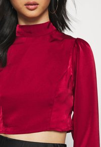 Burgundský saténový cropped top s dlouhými rukávy, vysokým límcem a s detaily na ramenech. Hladká textura a přiléhavý střih.