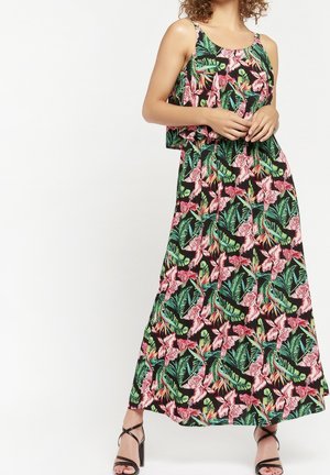 Femme portant une robe noire sans manches, longue jusqu'aux chevilles, avec des motifs floraux roses et feuillages verts, debout avec les mains jointes et portant des talons noirs.