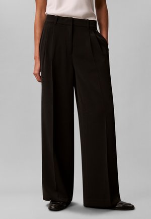 Woman wearing high-waisted, wide-leg black trousers with pleats and black shoes, standing against a plain light gray background.