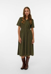 Vestido abotonado verde oliva hecho de una tela suave, con mangas cortas, cintura elástica y una falda fluida, combinado con botas marrones hasta la rodilla.