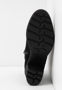 Bottine noire avec semelle plate en caoutchouc présentant un motif de bande de roulement en zigzag, tige en daim, fermeture éclair latérale et logo de la marque embossé sur le talon.