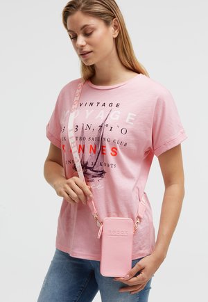 Junge Frau trägt ein rosa bedrucktes T-Shirt und blaue Jeans und hält eine hellrosa Umhängetasche für das Handy mit passendem verstellbarem Riemen.