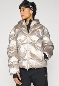 Metaliczna srebrna kurtka puchowa z teksturowanym pikowanym wzorem, przednim zamkiem błyskawicznym i kapturem. Zestawiona z czarnymi goglami narciarskimi i ciemnymi spodniami.