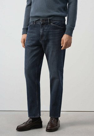 Homme portant un jean droit bleu foncé avec les ourlets roulés, des chaussettes noires, des mocassins en cuir marron et un pull bleu à manches longues, debout sur un sol gris.