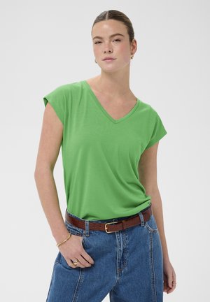 Jeune femme portant un haut vert à manches courtes avec col en V, rentré dans un jean bleu taille haute avec une ceinture marron, sur fond blanc.