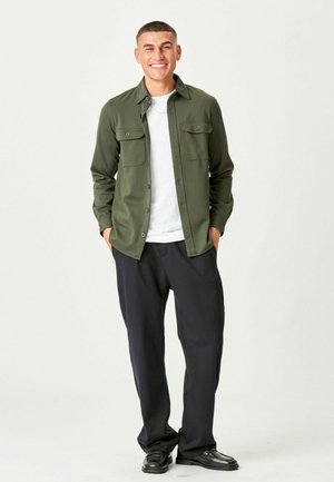 Lächelnder Mann mit kurzen Haaren, der ein olivgrünes Hemd über einem weißen T-Shirt, schwarze Hose und schwarze Schuhe trägt, die Hände in den Taschen.