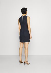 Žena s krátkými tmavými vlasy stojí zády, oblečená v bezramínkových námořnických modrých šatech a černých sandálech na vysokém podpatku s páskem kolem kotníku.