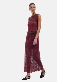 Set di due pezzi all'uncinetto in color bordeaux composto da un top corto con design a scallop e una lunga gonna trasparente con dettagli a pattern. Sandali neri.