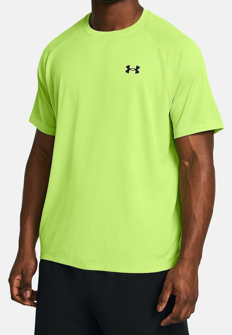 Man i en ljus limegrön kortärmad Under Armour träningströja och svarta shorts, som står mot en enkel ljus bakgrund.
