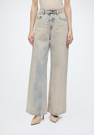 Donna che indossa jeans beige scoloriti a vita alta e gamba larga con una leggera tinta blu, abbinati a décolleté a punta color nude e top beige senza maniche.