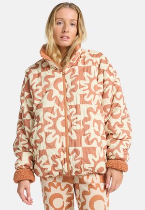 Woman wearing a quilted jacket and pants with matching abstract wavy orange and cream floral patterns and fleece-lined cuffs.