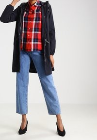 Osoba v černé bundě s kapucí na červenou kostkovanou košili, světle modrých džínech a černých lodičkách, stojí s jednou rukou zvednutou.