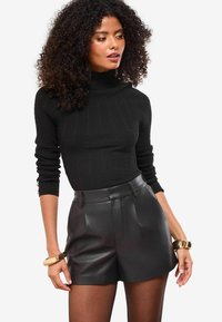 Sieviete ar cirtainiem matiem, ģērbta melnā ribotā augstajā kaklā svārkā, melnās ādas šortos, caurspīdīgā zeķēs un zeltainajās rokassprādzēs, pozē ar atslābinātiem palīdz.