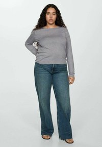 Siva dug jakna dugih rukava uparena s lepršavim plavim trapericama. Traperice imaju izrezan rub i širok kroj. Model stoji naslonjen na svijetlu pozadinu.