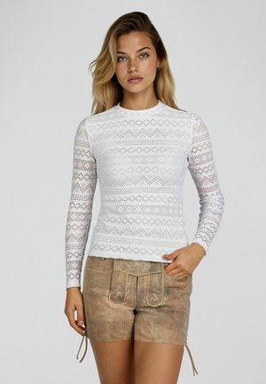 Frau trägt weißes Spitzen-Longsleeve und braune Lederhose im traditionellen Stil, steht vor einfarbigem hellgrauem Hintergrund.