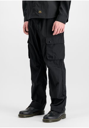 Person trägt schwarze Cargo-Hosen mit großen Seitentaschen und schwarzen Lederschuhen, steht vor weißem Hintergrund.