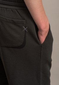 Jogging grigi scuri con tasca a toppa, caratterizzati da una leggera texture e un dettaglio decorativo in grigio a punto croce sulla tasca.