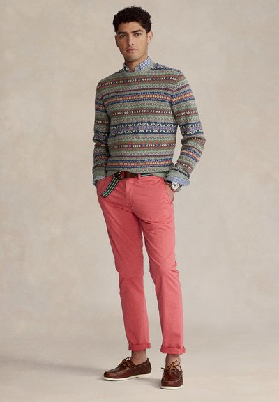 Πλεκτό πουλόβερ σε πράσινο χρώμα με πολύχρωμα γεωμετρικά σχέδια, συνδυασμένο με παντελόνι κοραλί και καφέ παπούτσια τύπου boat. Δι αθέτει γιακά με κουμπιά και μανσέτες που γυρίζουν.