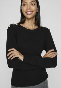 Pull en tricot noir avec texture côtelée, col rond et quatre boutons décoratifs dorés à l'épaule. Manches longues avec poignets ajustés.
