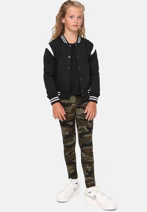 Veste bomber noire avec des rayures blanches, haut noir et leggings camouflage. Des baskets blanches complètent la tenue. Textures lisses et coupe décontractée.