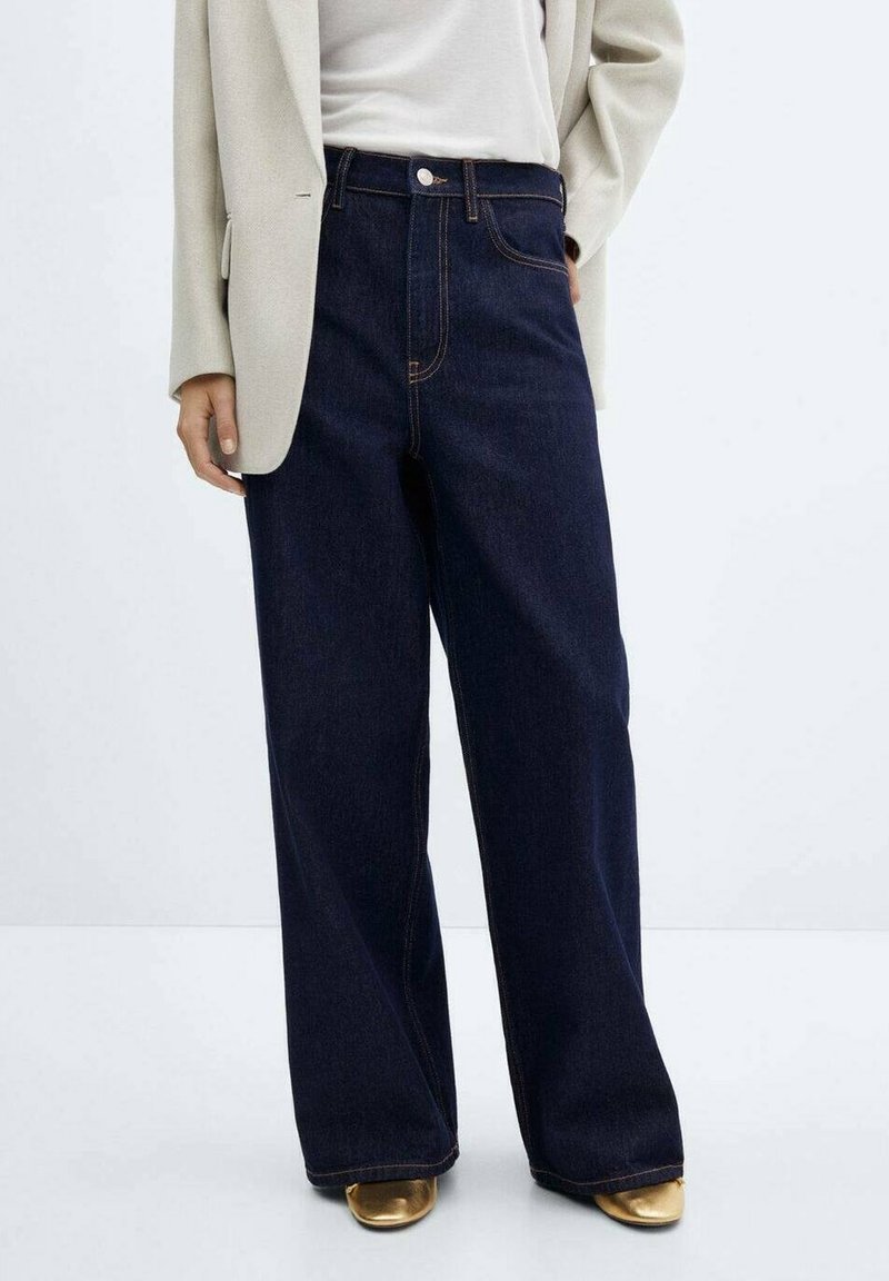 Personne portant un jean en denim sombre à taille haute et jambes larges, un blazer beige, un haut blanc et des chaussures dorées à bout pointu, debout devant un fond uni.
