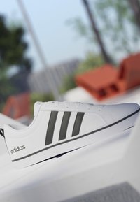 Zapatilla blanca de Adidas con tres franjas negras y logotipo en el lateral, colocada sobre una superficie blanca con fondo exterior desenfocado.